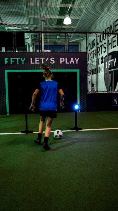 Person in a blue shirt standing on an indoor soccer field kicking ball through acura elite wireless circuit