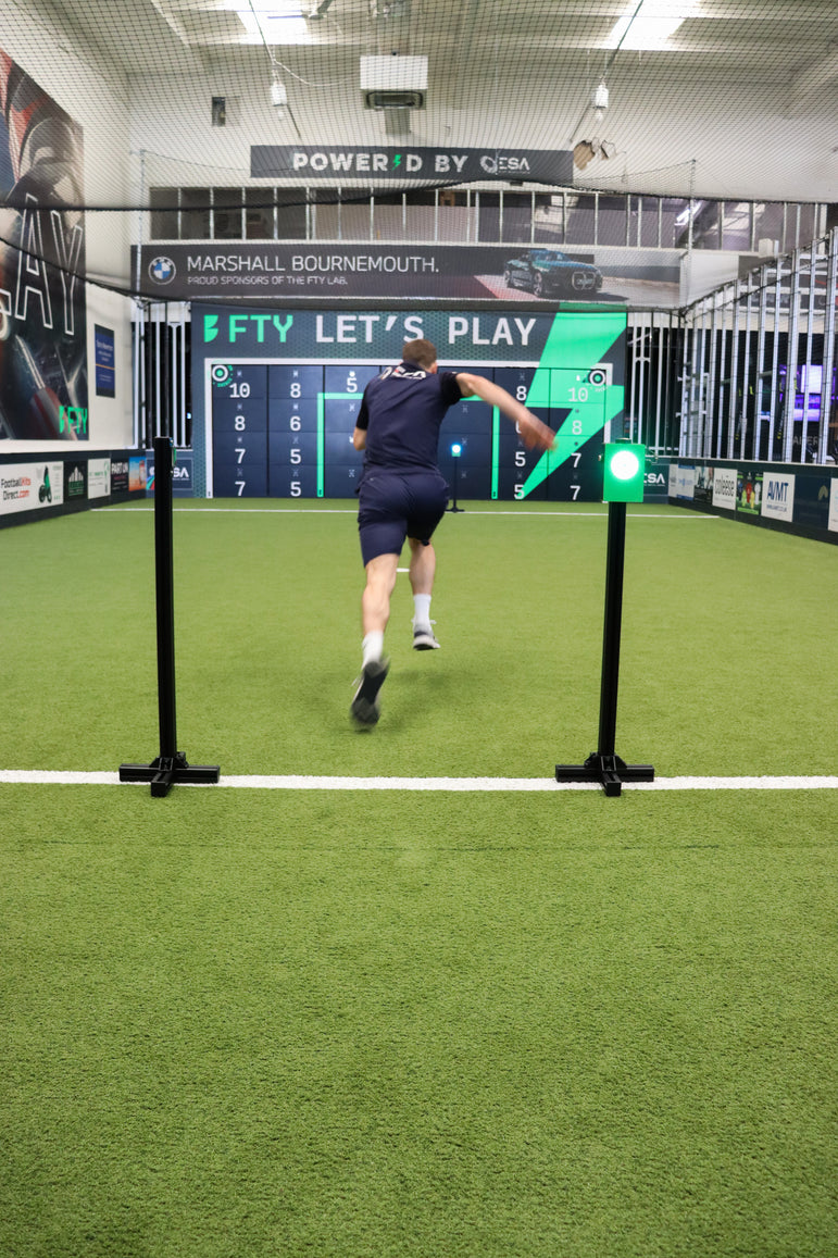 A man in a dark navy athletic outfit runs across a green turf indoor driving range, approaching an acura elite precision wall 28 panel while running through a wireless sprint 