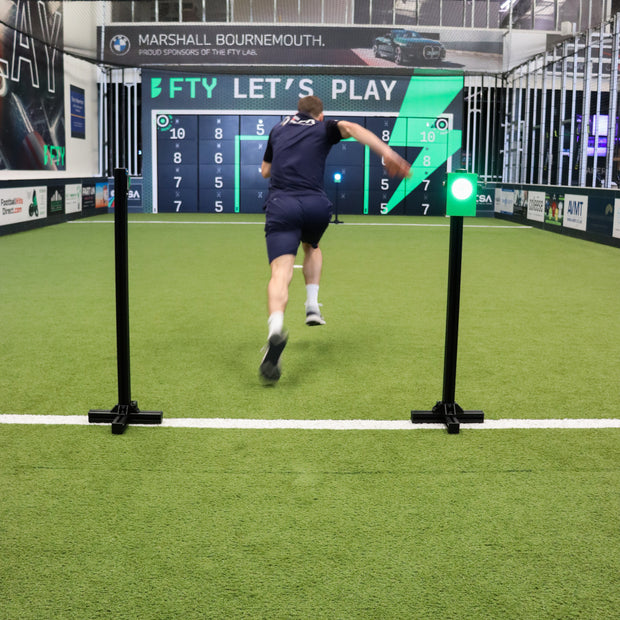 A man in a dark navy athletic outfit runs across a green turf indoor driving range, approaching an acura elite precision wall 28 panel while running through a wireless sprint 