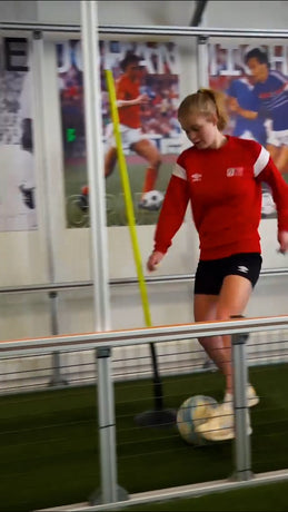 A woman in a red long-sleeve athletic top and black shorts dribbling a soccer ball on an indoor turf field, with a yellow training pole and sports wall posters in the background using acura elite training lane (dribble)