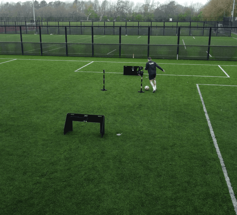 A person in a black long-sleeve top and light shorts runs on a green soccer field near acura elite wireless circuit and a black fence.