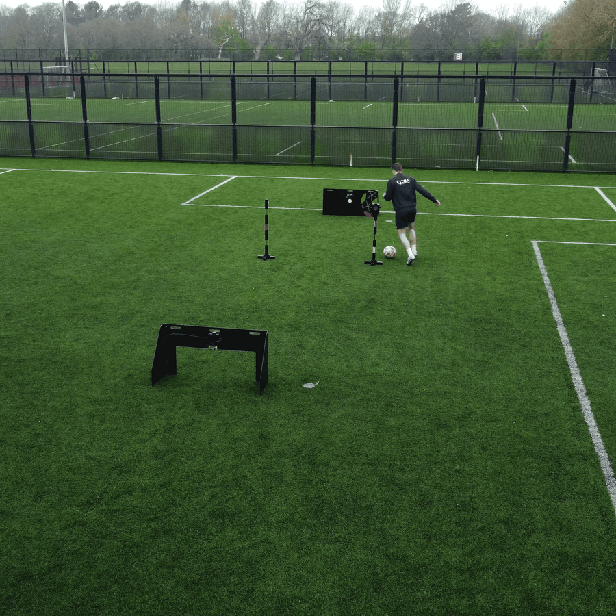 A person in a black long-sleeve top and light shorts runs on a green soccer field near acura elite wireless circuit and a black fence.