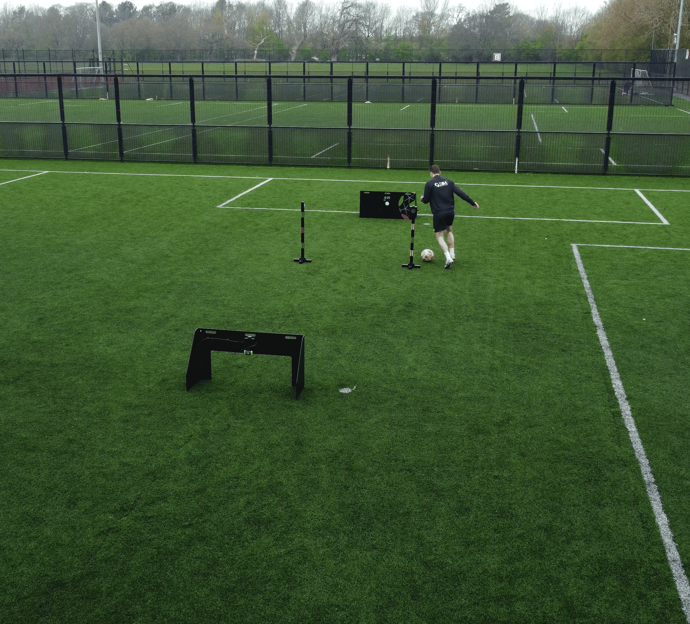A person in a black long-sleeve top and light shorts runs on a green soccer field near acura elite wireless circuit and a black fence.