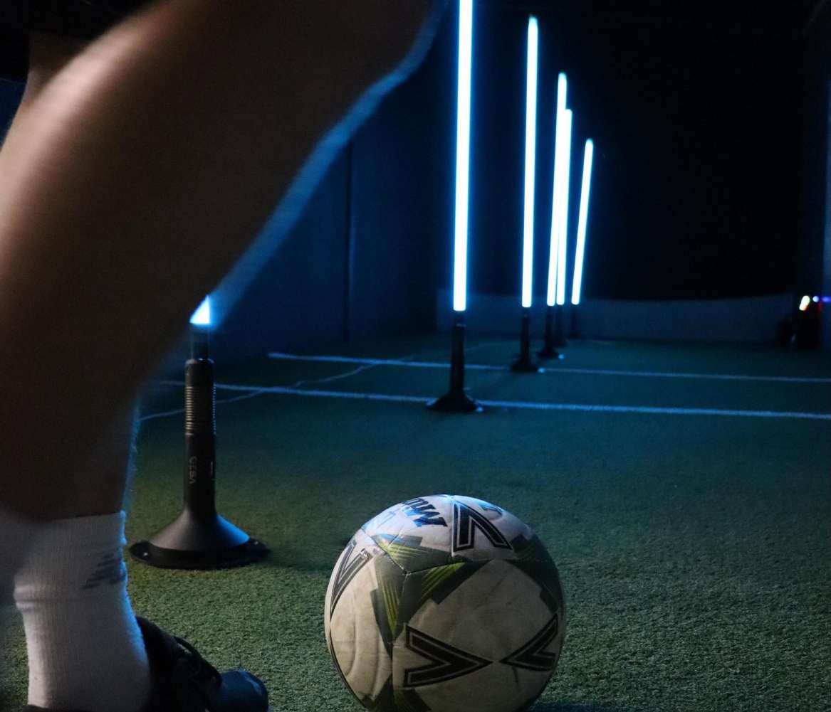 A soccer ball with geometric patterns sits on green turf in a dark indoor training facility, with a person's leg in the foreground and vertical blue LED light poles in the background from training lane (dribble)