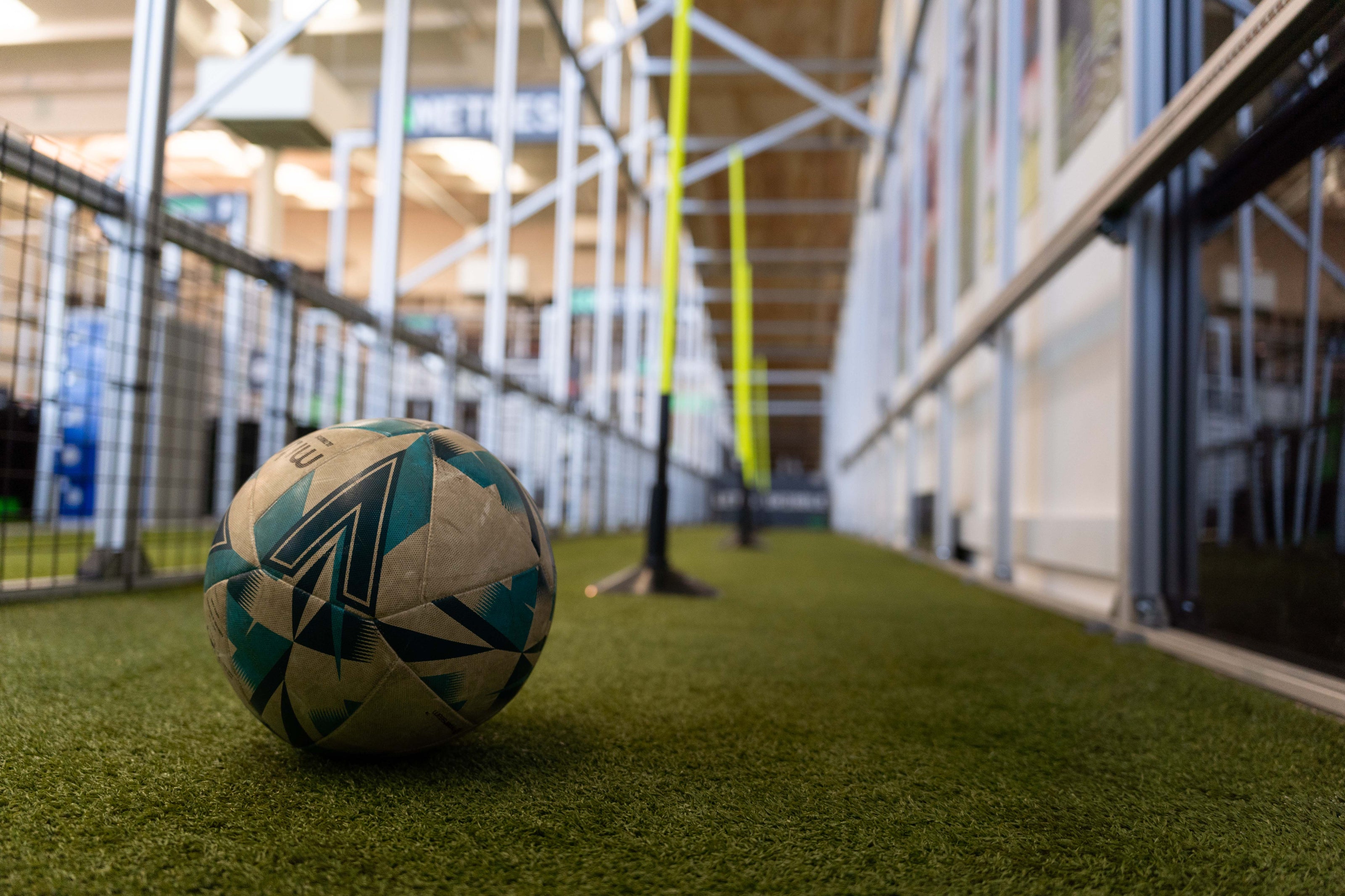 A soccer ball with a geometric pattern of white, blue, and black sits on artificial turf inside an indoor sports facility with a blurred background showing the acura elite training lane (dribble)