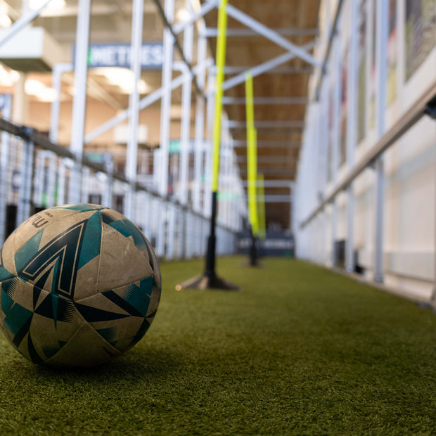A soccer ball with a geometric pattern of white, blue, and black sits on artificial turf inside an indoor sports facility with a blurred background showing the acura elite training lane (dribble)