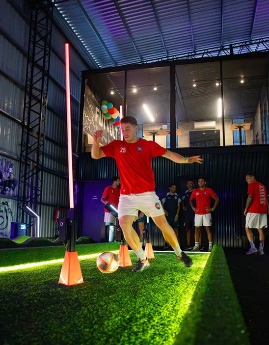 A man in a red t-shirt and white shorts kicks a soccer ball in an indoor sports facility with artificial turf, LED light poles, and other players in the background using acura elite training lane (dribble)