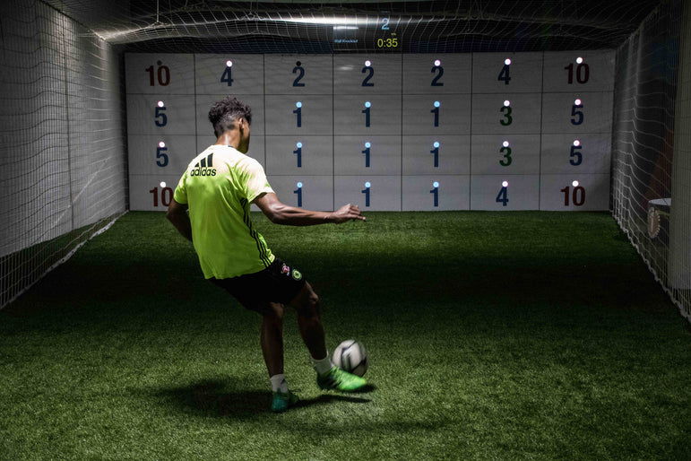 man in football kit kicking ball at acura elite 28-panel precision wall with number scores on panels