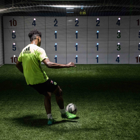 man in football kit kicking ball at acura elite 28-panel precision wall with number scores on panels