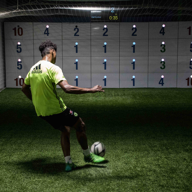 man in football kit kicking ball at acura elite 28-panel precision wall with number scores on panels