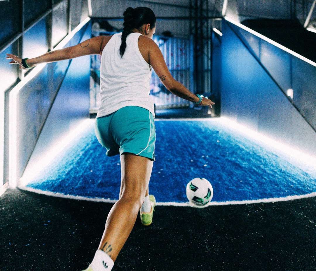 A woman in a white tank top and teal shorts kicks a soccer ball on a blue illuminated indoor court at acura elite esa power shot