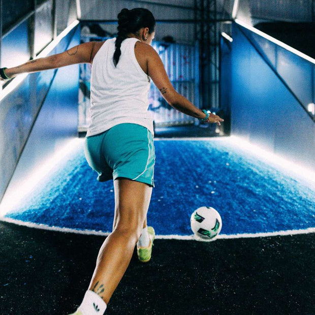 A woman in a white tank top and teal shorts kicks a soccer ball on a blue illuminated indoor court at acura elite esa power shot