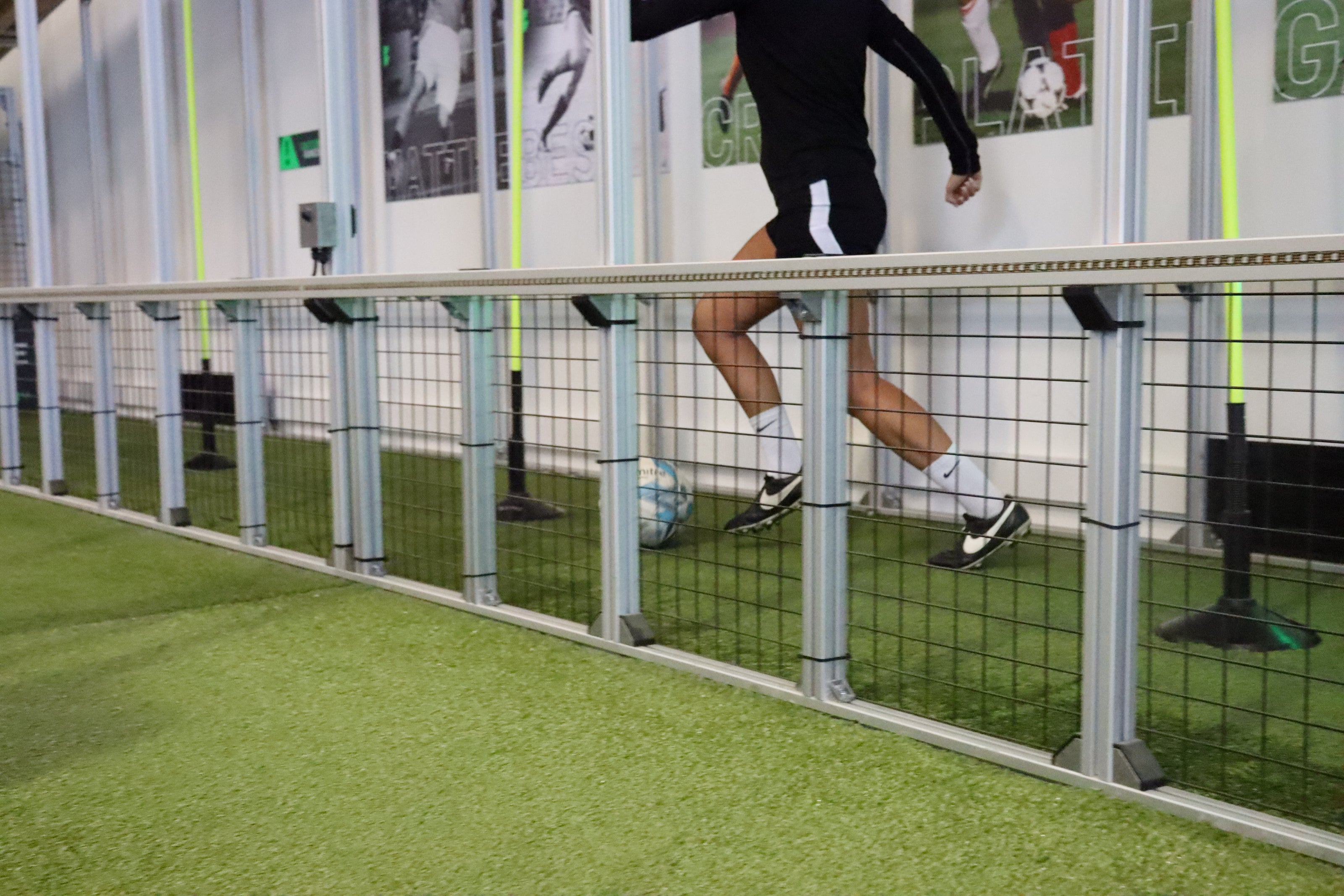 A person in black athletic clothing runs along an indoor training lane (dribble) with a silver and black wire mesh barrier, with green artificial turf flooring and wall posters in the background.