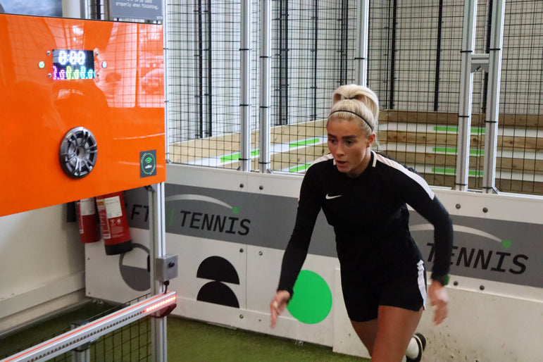 A woman in a black athletic long-sleeve top and shorts with white side stripes is running indoors on acura elite training lane (sprint)