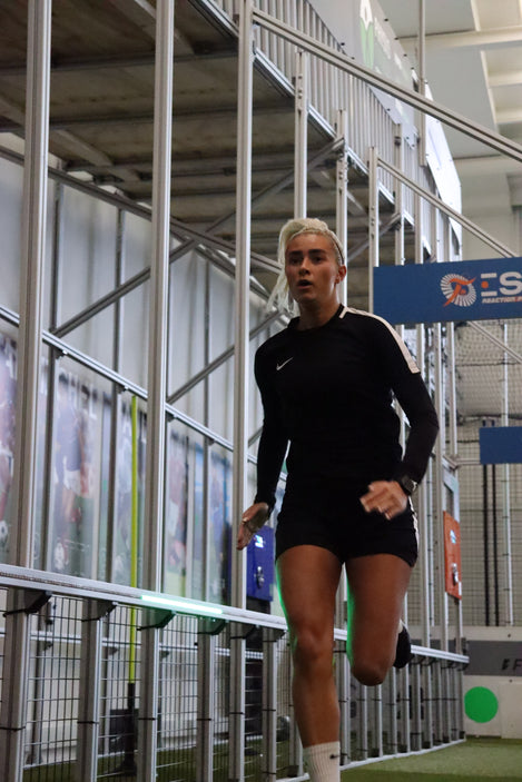 A person in a black athletic outfit with white trim is running indoors on a sprint lane surrounded by metal fencing and banners.